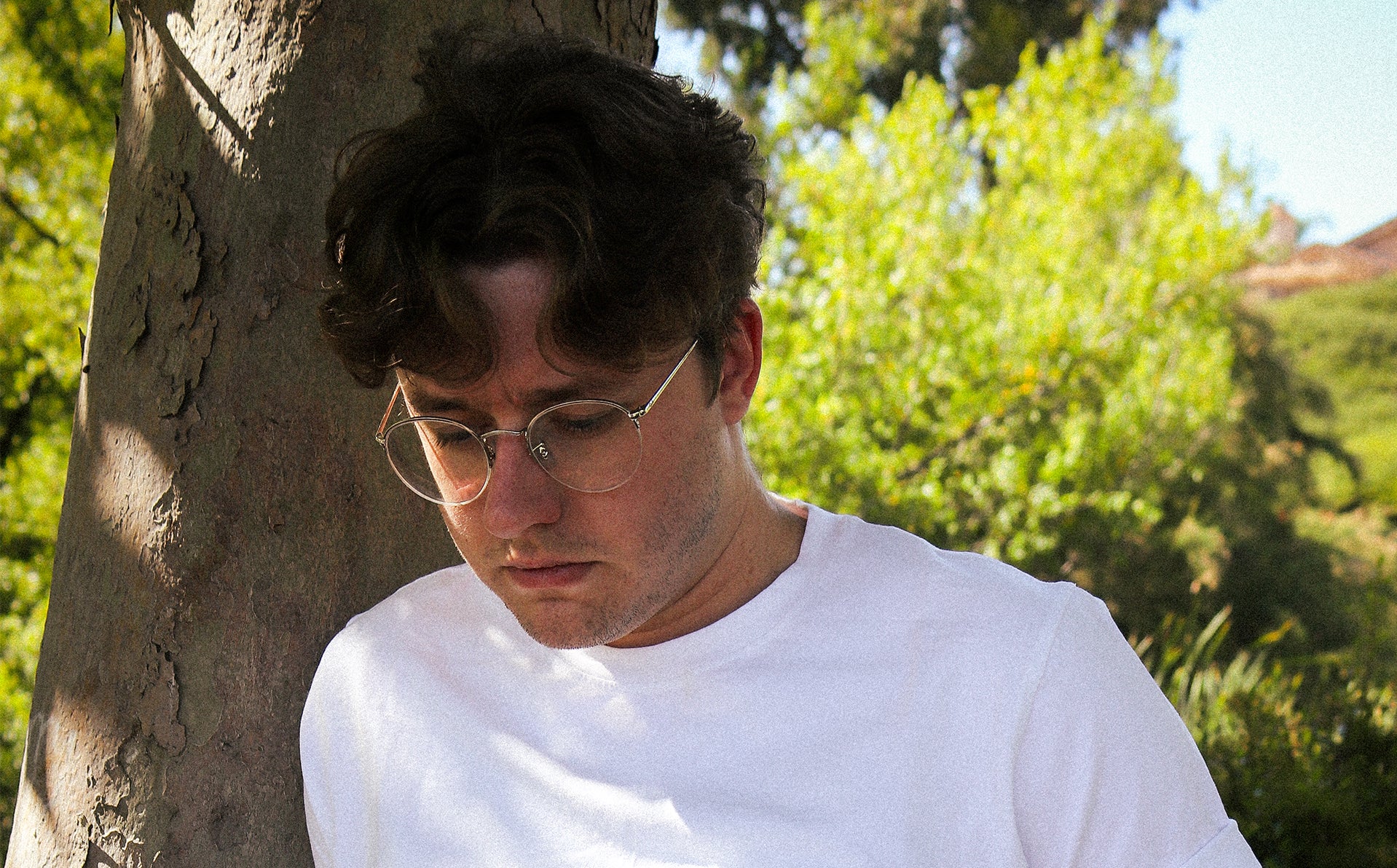 Model leaning on tree wearing white t-shirt with lush green foliage in background