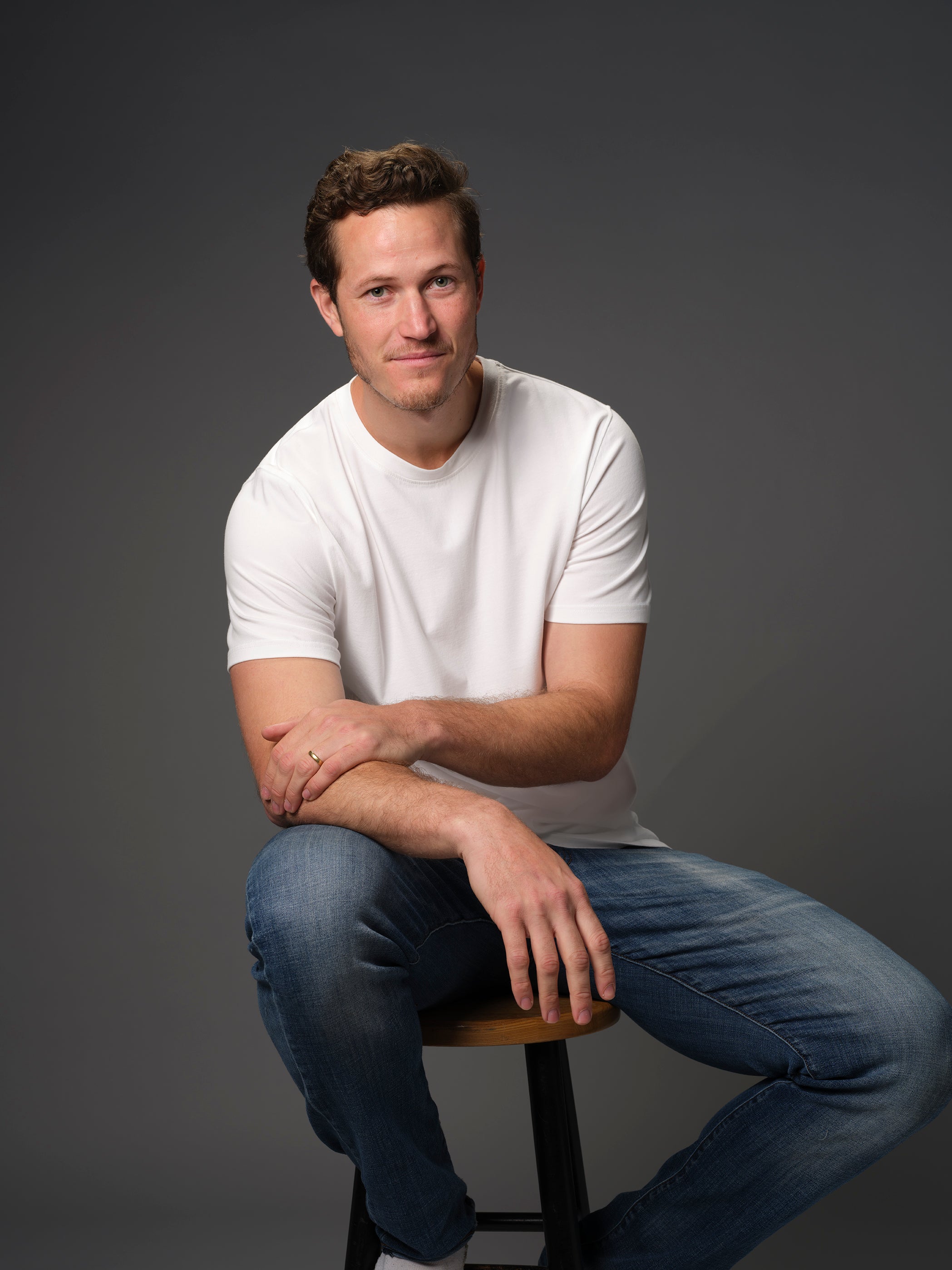 Male model with slight grin seated on stool wearing our white t-shirt dark background