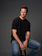 Seated male model grinning wearing our premium black t-shirt dark background