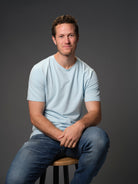 Male model with small grin seated on stool wearing our "breakwater" light blue t-shirt