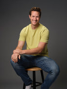 Male model smiling seated on stool wearing our “light moss” green t-shirt dark background