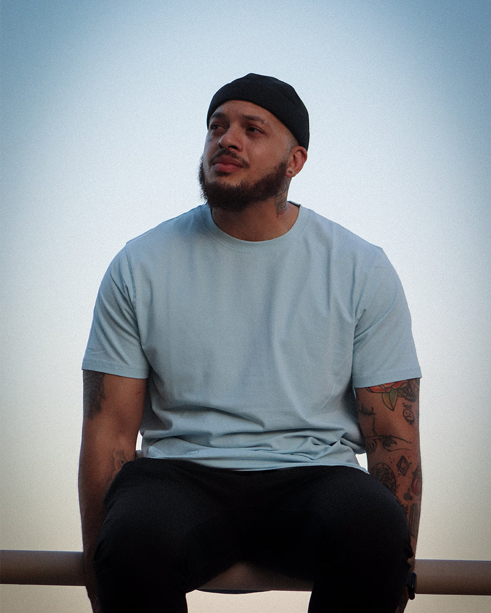 Tattooed model sitting on railing in light blue t-shirt with clear blue sky in background